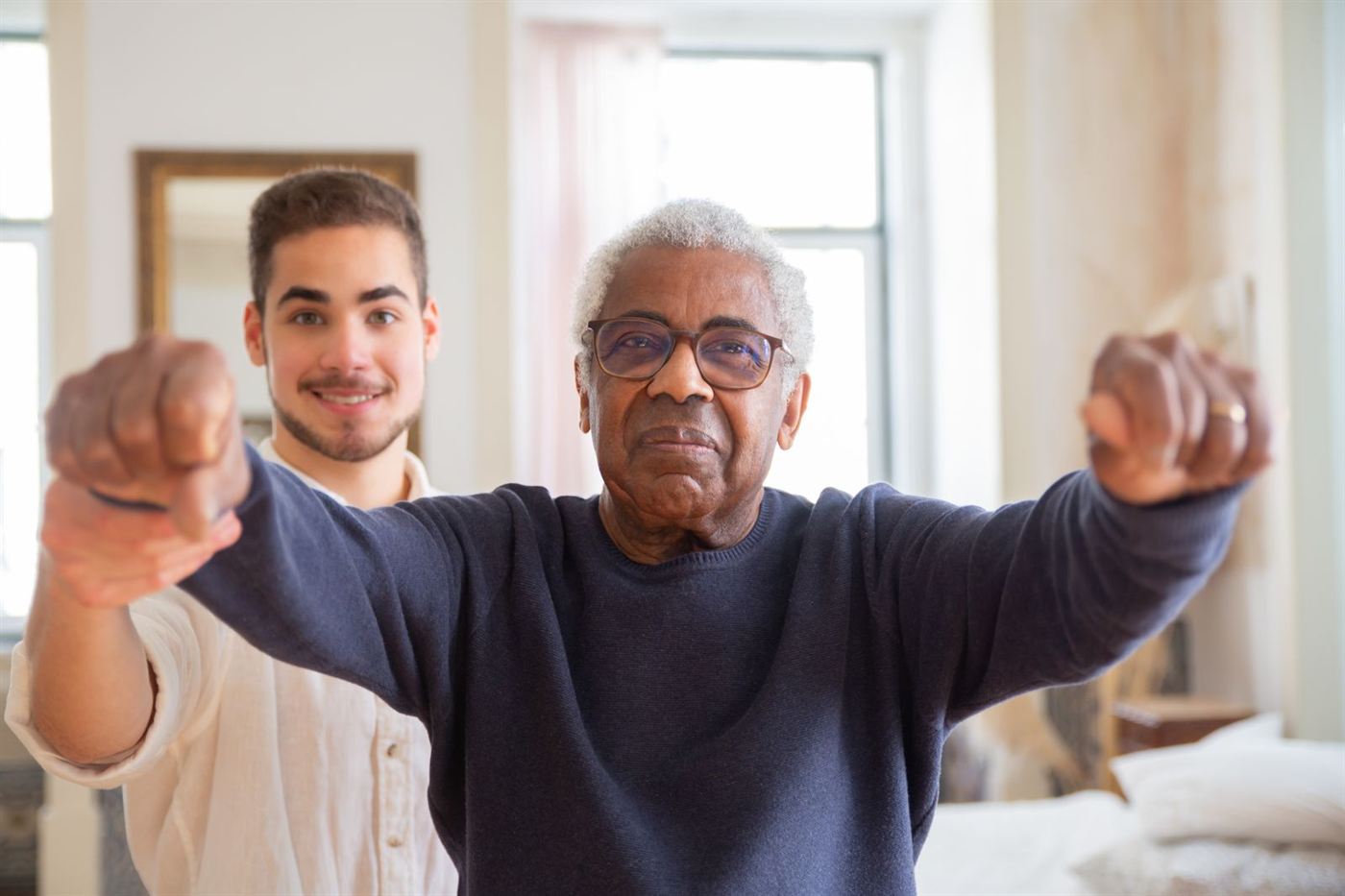 A senior man practicing a balance exercise with his arms extended while a young trainer guides him